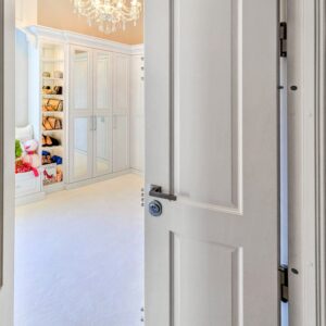 Elegant white panic room door with 10 steel bolts extended, leading to white walk in closet with purses and shoes.