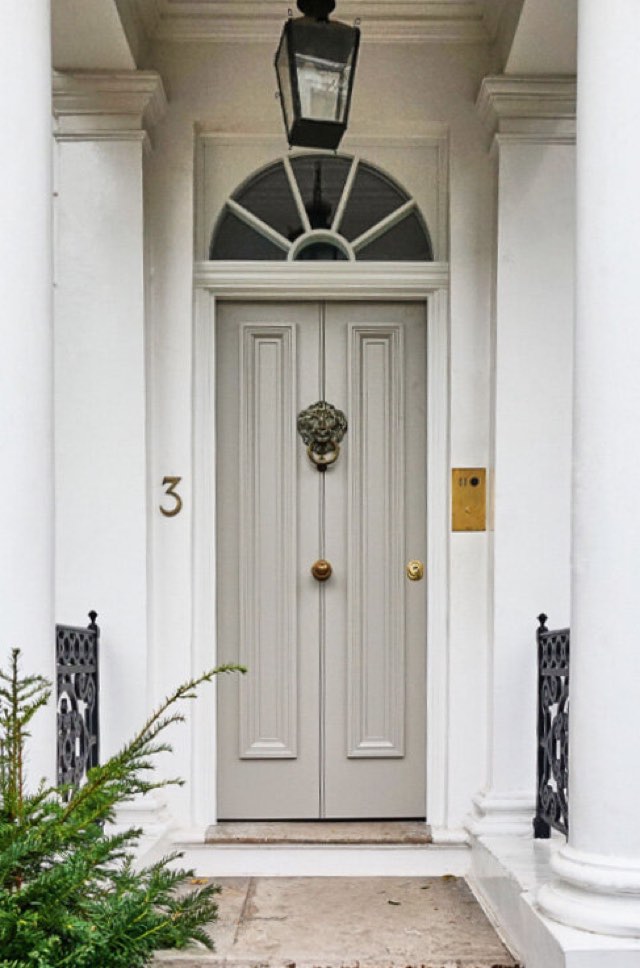 Bulletproof front door in a white stucco portico — a pale panelled door with a brass lion knocker, fanlight above, giving no sign of the ballistic steel inside.