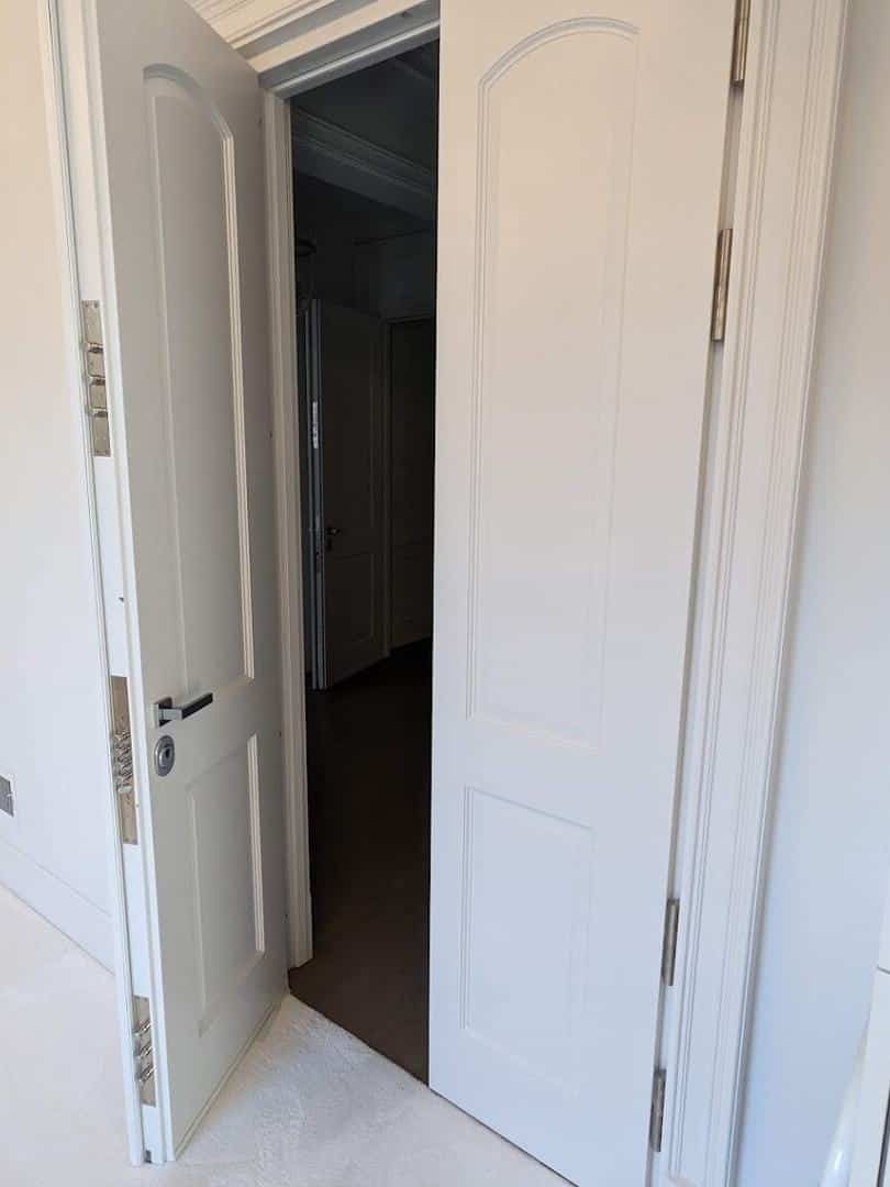 White paneled armored double door standing open onto a dark hallway, steel bolts in the edge of the left leaf.